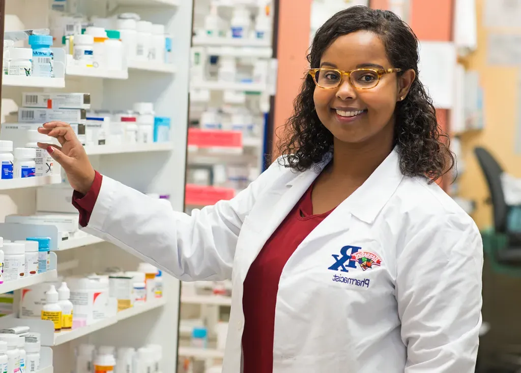 A pharmacy student smiles as she reaches for a bottle of medication from a large shelving unit